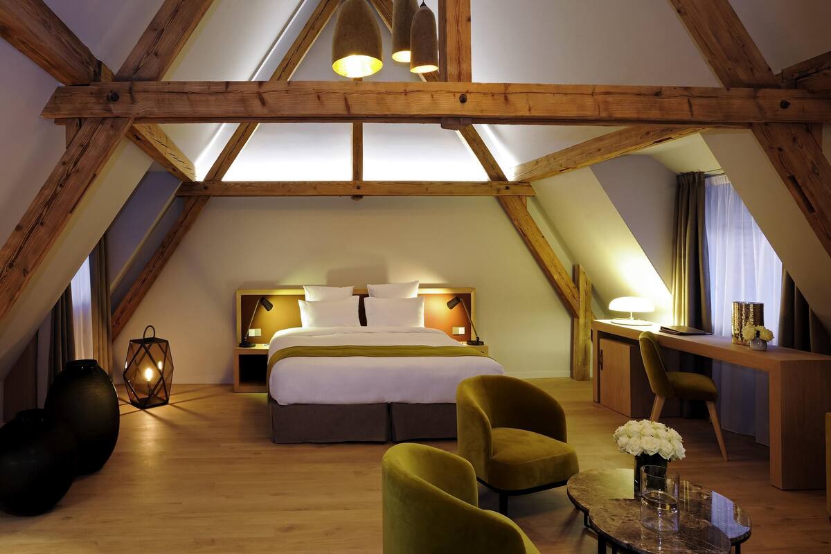 A high-ceilinged room with a bed, two chairs, a desk, and a lamp.