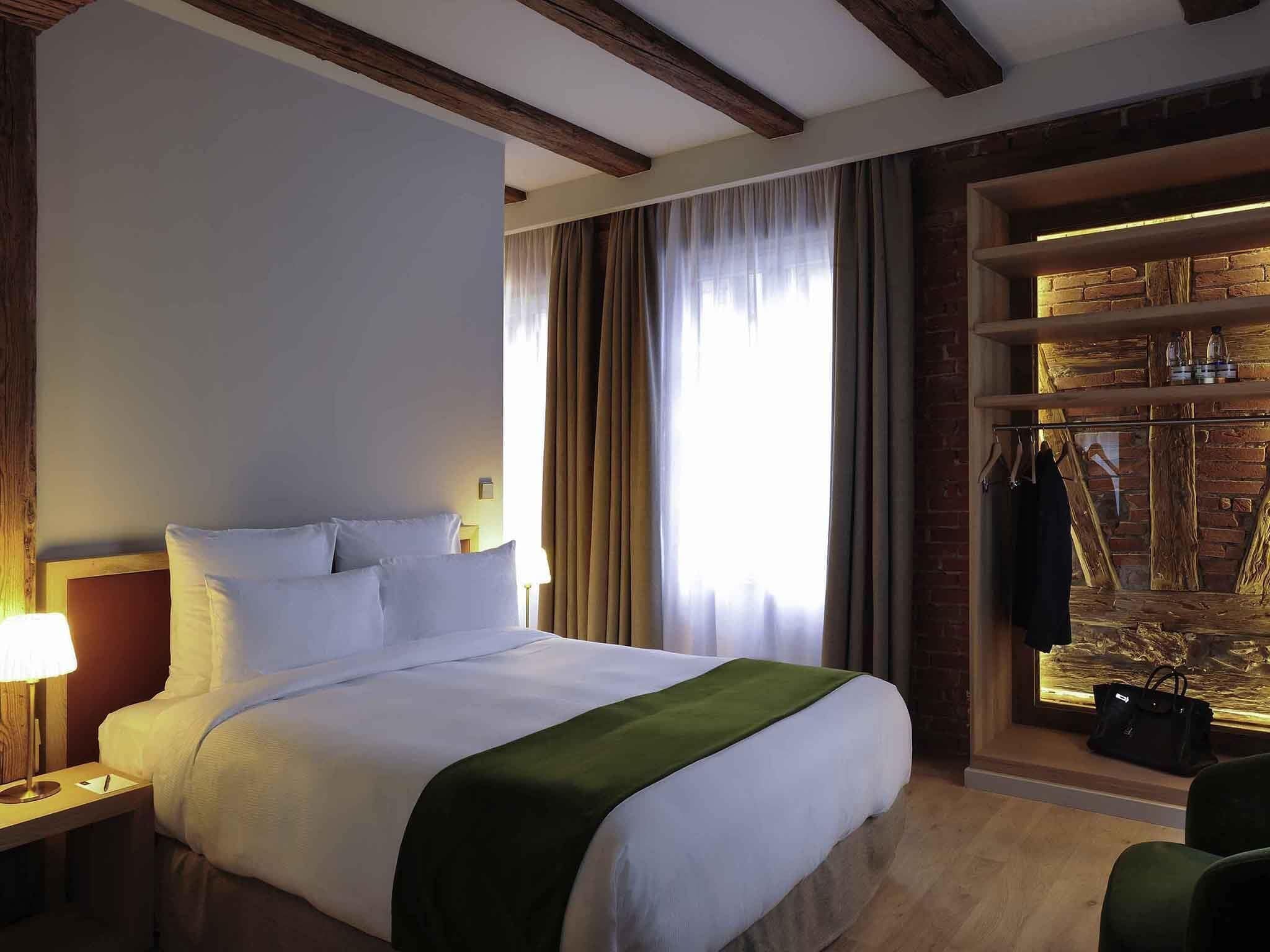 Ein Hotelzimmer mit einem Bett, einem Nachttisch, einer Lampe, einem Fenster mit Vorhängen und einer Ziegelwand mit Ablage.