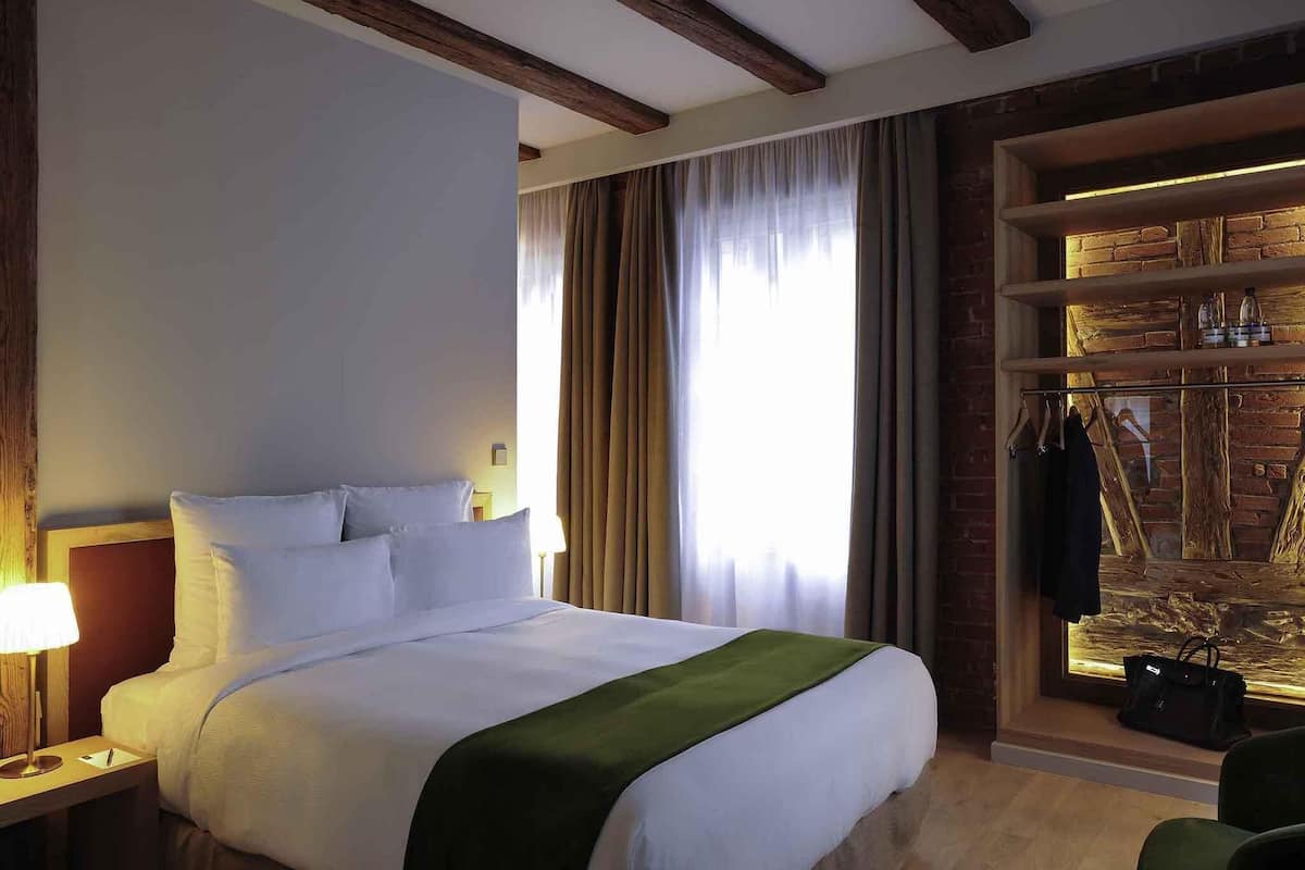 A hotel room with a bed, a nightstand, a lamp, a window with curtains, and a brick wall with a shelf.