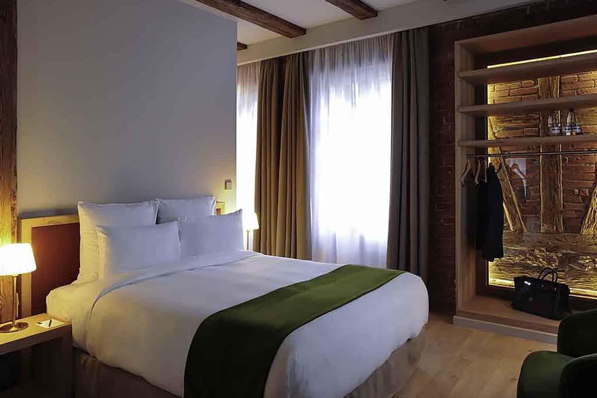 A hotel room with a bed, a nightstand, a lamp, a window with curtains, and a wooden wall with a coat rack.