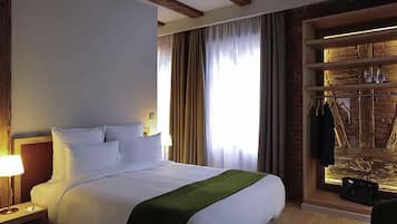 Une chambre d’hôtel comprenant un lit, une table de chevet, une lampe, une fenêtre avec des rideaux et un mur en bois muni d’un porte-manteau.