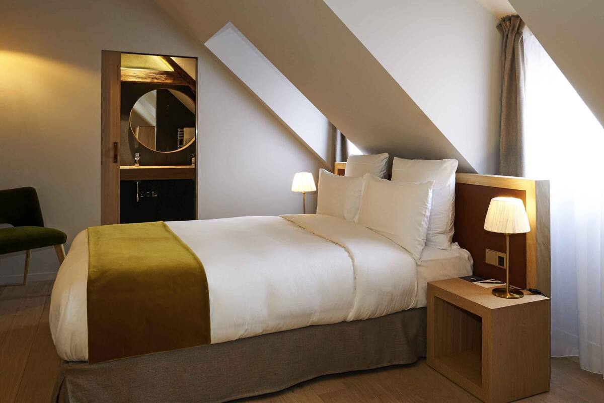A modern hotel room with a large bed, a bedside table with a lamp, a mirror, and a green chair.
