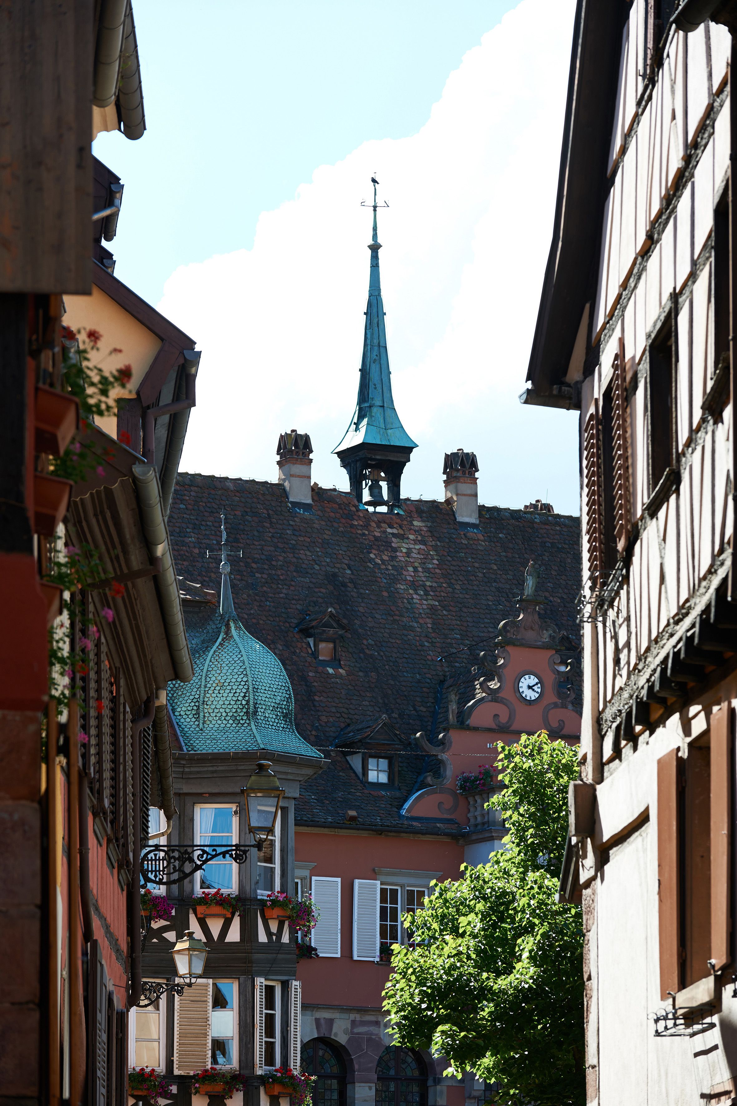 A narrow street with traditional European architecture, including a church steeple and a building with a clock.