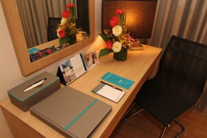 A hotel desk with a mirror, a vase of flowers, a calendar, a pen, and a notebook.