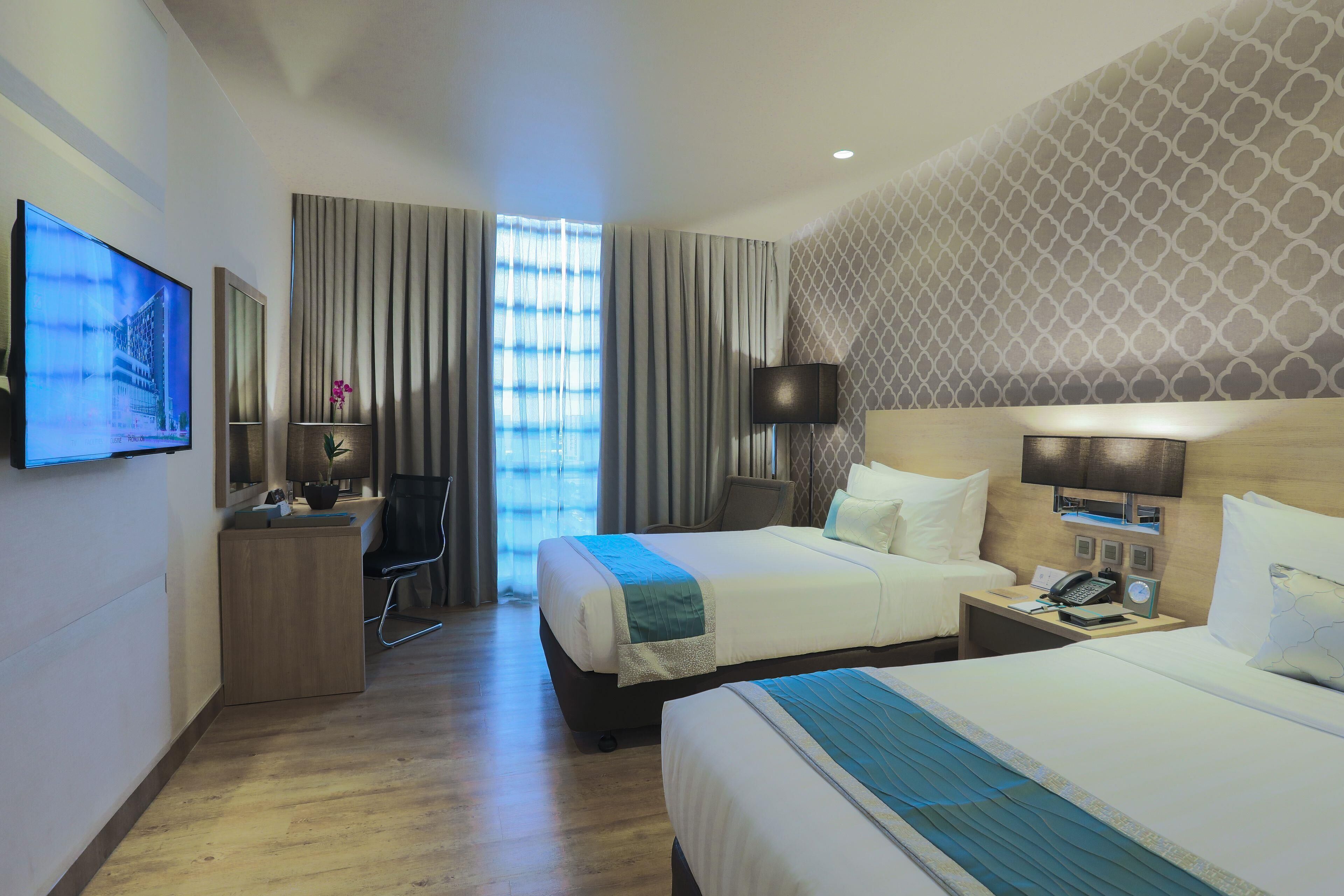 A hotel room with two beds, a desk, a TV, and a window with curtains.