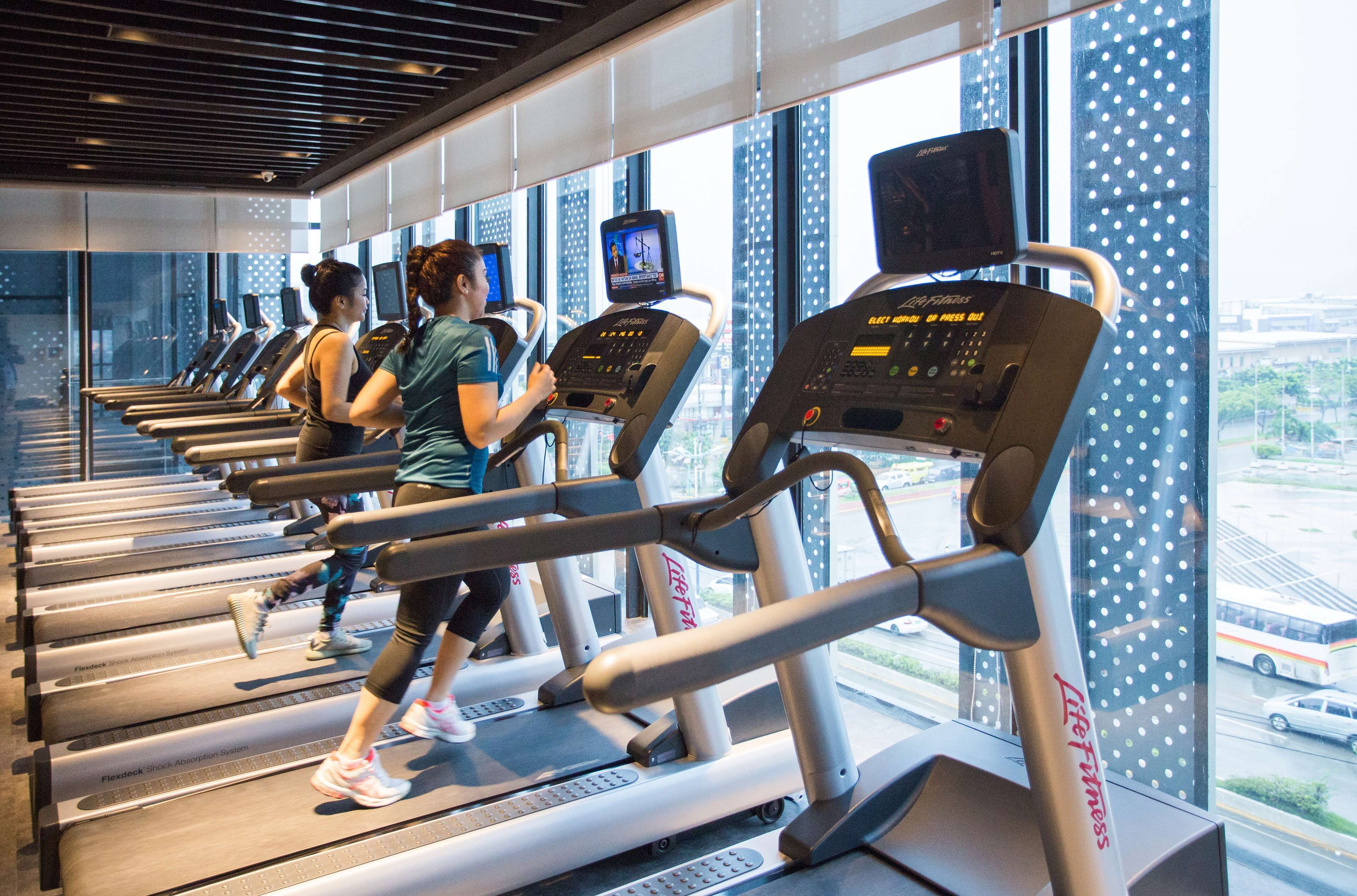 Une salle de sport équipée de tapis de course, d’appareils d’exercice et de grandes fenêtres donnant sur la ville.