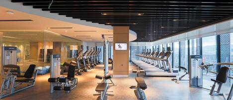 A modern gym with various exercise equipment, large windows, and a high ceiling.
