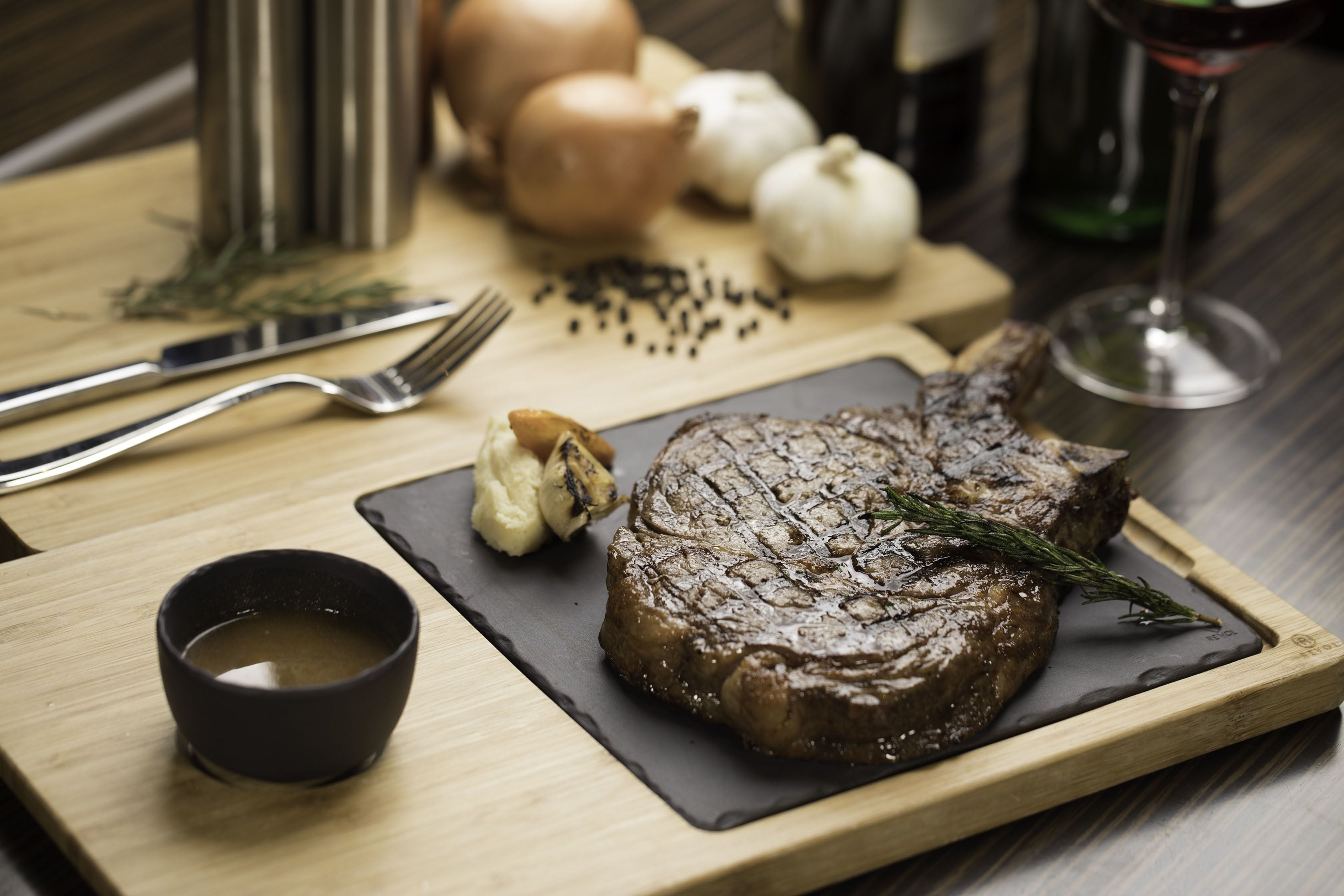 Un bistec con ajo y romero sobre una tabla de madera, acompañado de un recipiente pequeño con salsa y una copa de vino tinto.