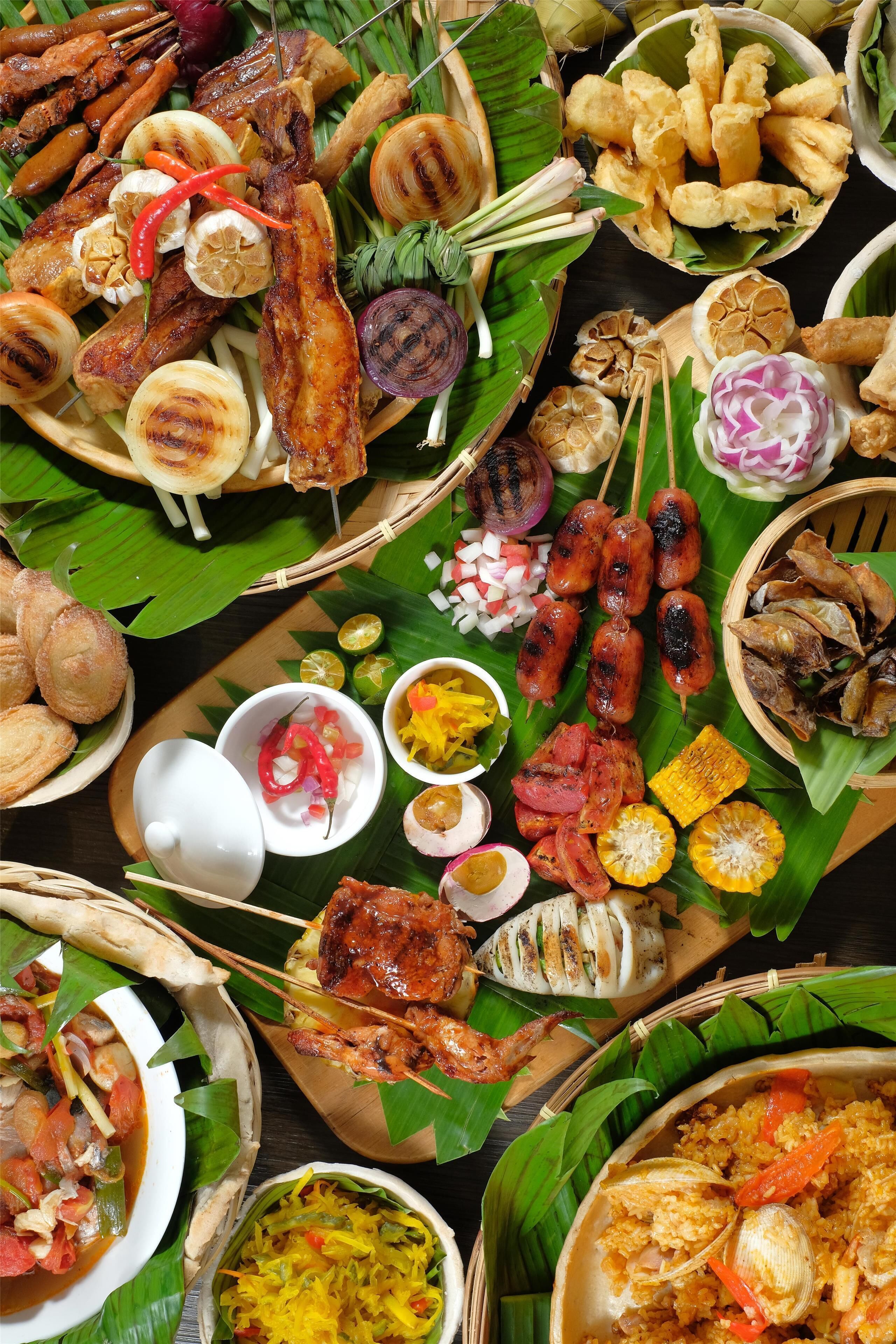 Variedad de carnes a la parrilla, verduras y guarniciones dispuestas sobre una mesa cubierta con hojas de plátano.