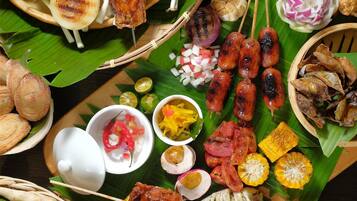 Variedad de carnes a la parrilla, verduras y guarniciones dispuestas sobre una mesa cubierta con hojas de plátano.