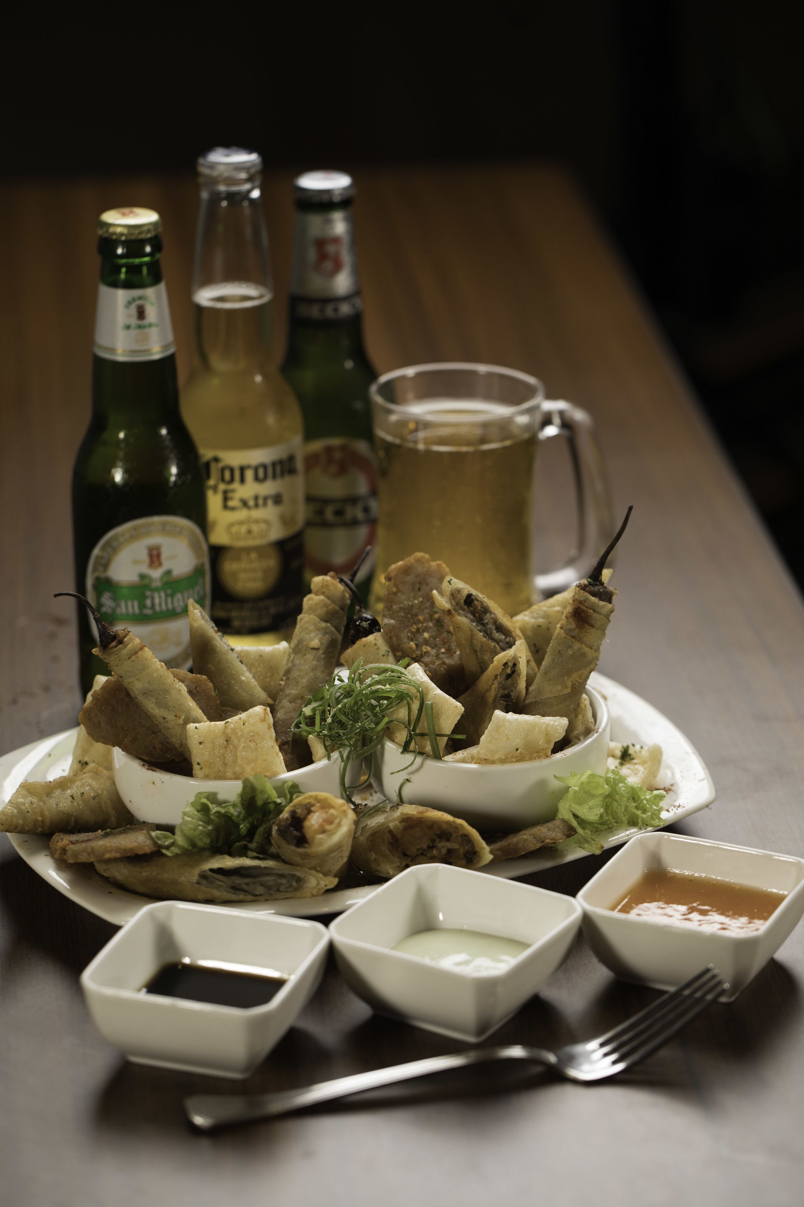 A plate of fried snacks with dipping sauces, a beer mug, and bottles of beer on a table.
