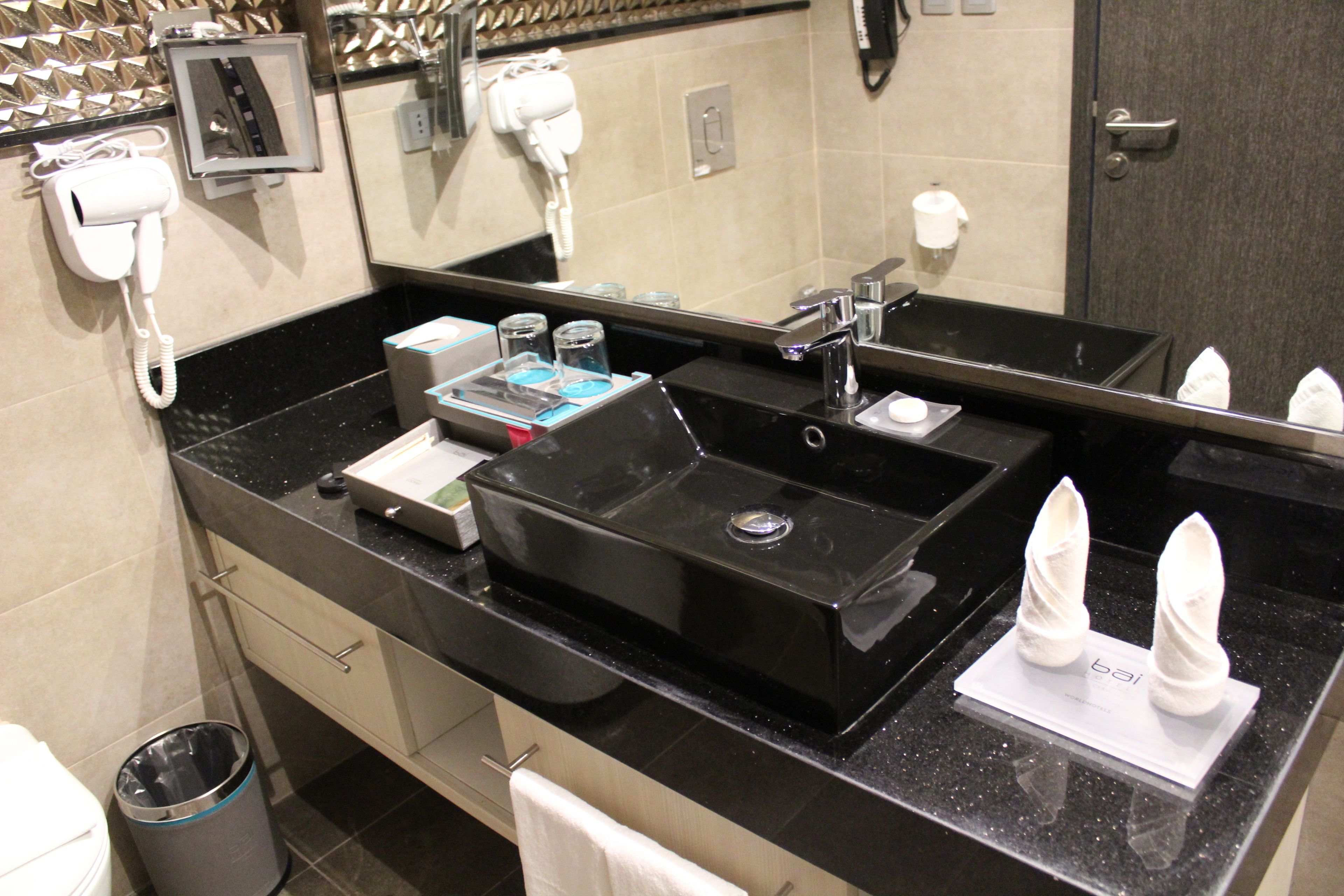 Un baño moderno con un lavamanos negro, un espejo, un secador de pelo y un cubo de basura.