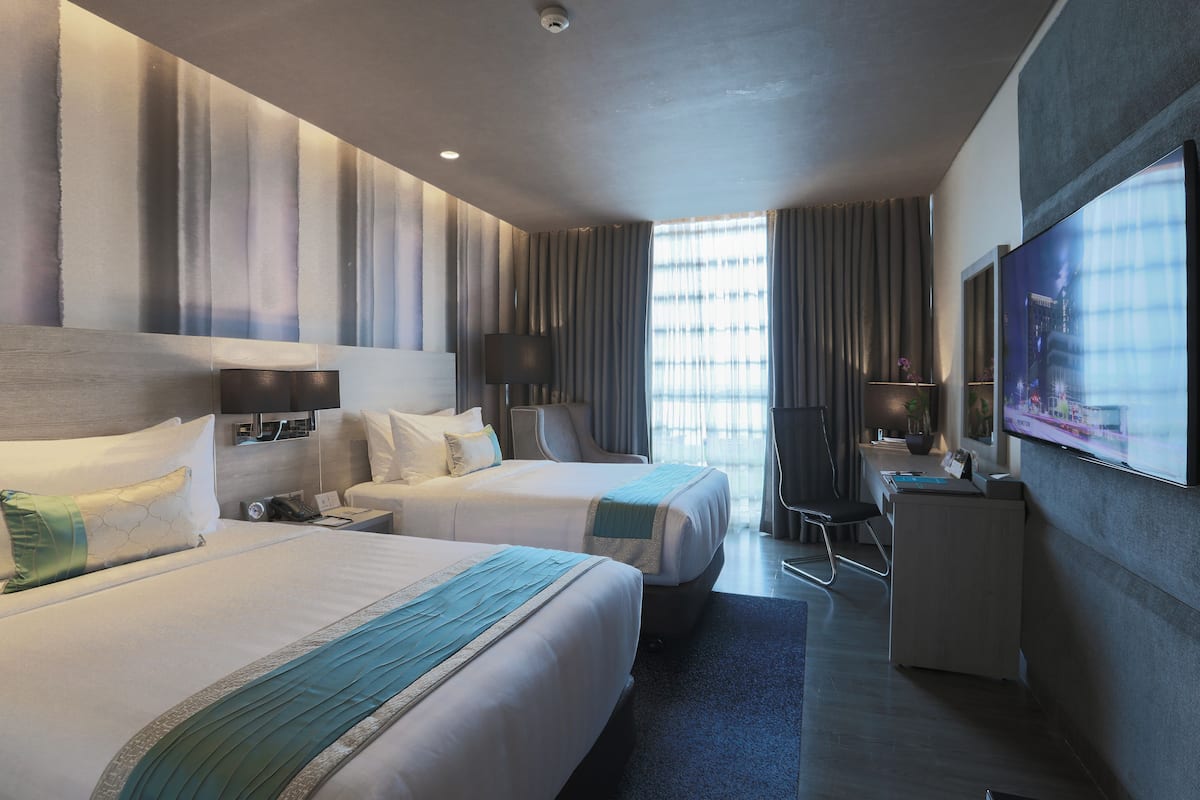 A hotel room with two beds, a desk, a chair, a TV, and a large window with curtains.