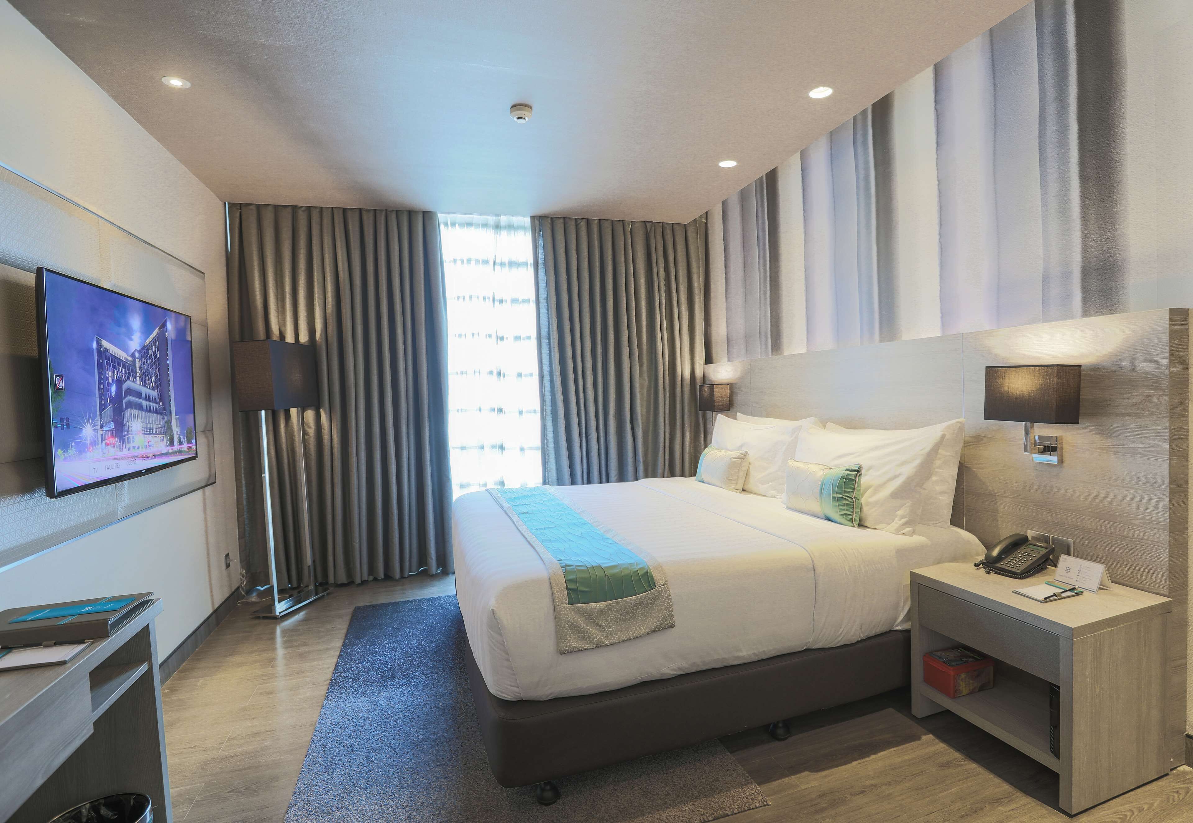 A modern hotel room with a large bed, a flat-screen TV mounted on the wall, a bedside table with a lamp, and a nightstand with a phone and a notepad.