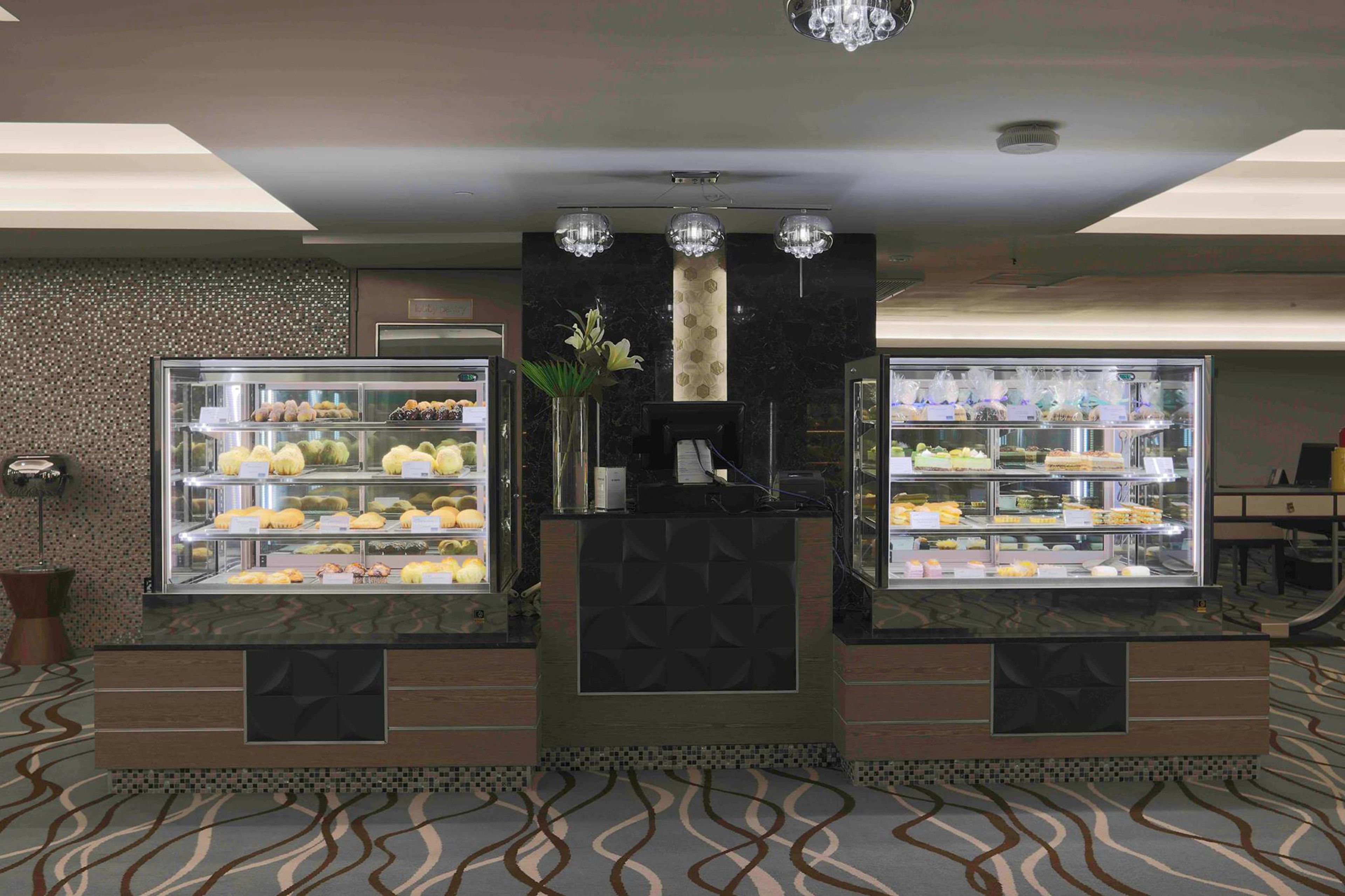 A hotel lobby with two glass display cases filled with pastries and fruits, a central decorative column, and patterned carpet flooring.