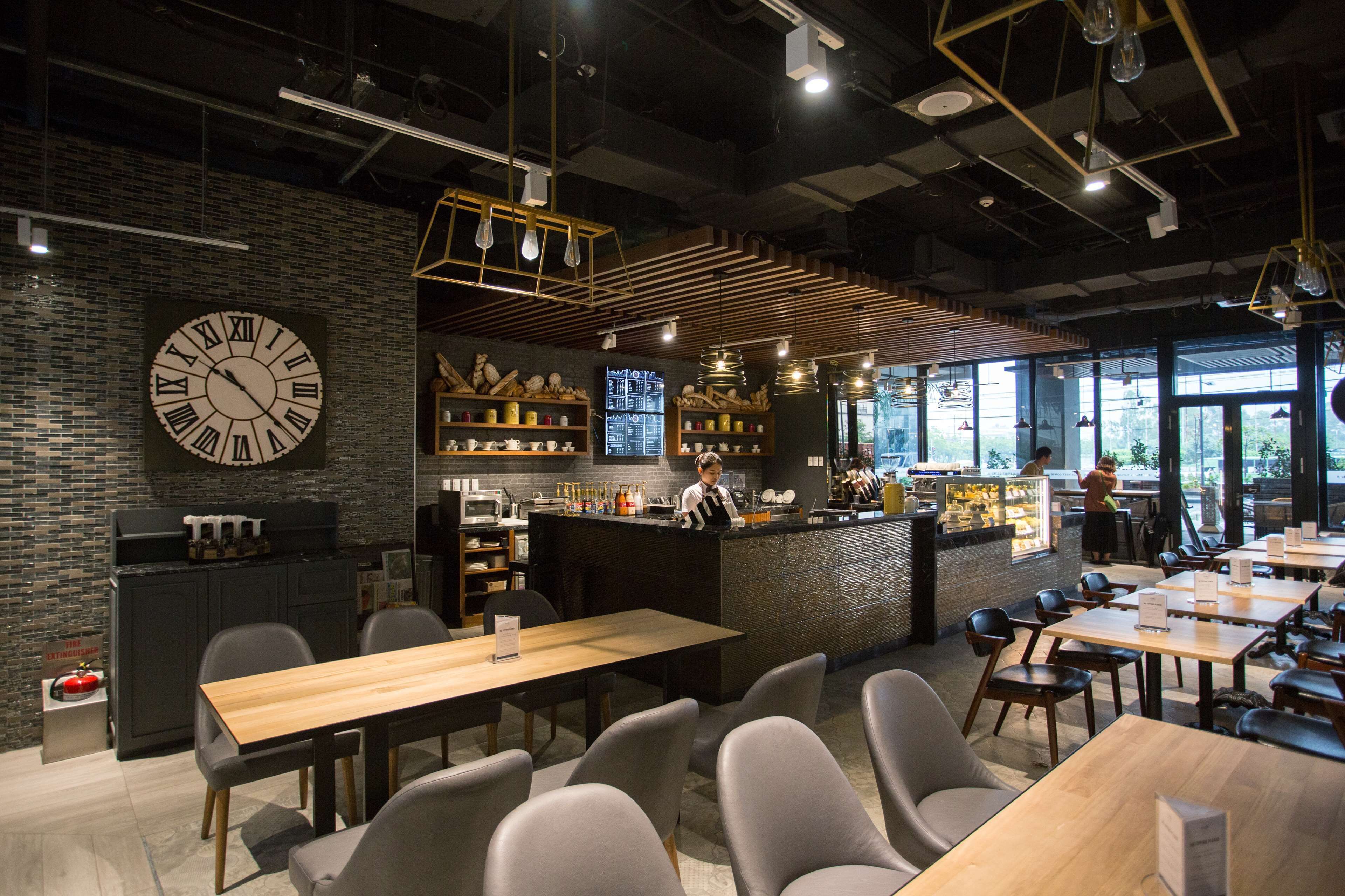 A modern cafe with a large wall clock, wooden tables, and chairs, and a bar area with a counter and shelves.