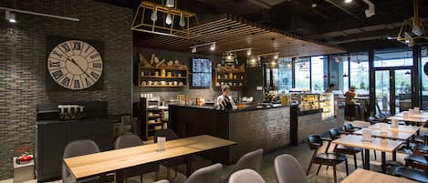 Una cafetería moderna con un gran reloj de pared, mesas y sillas de madera, y una zona de bar con mostrador y estanterías.