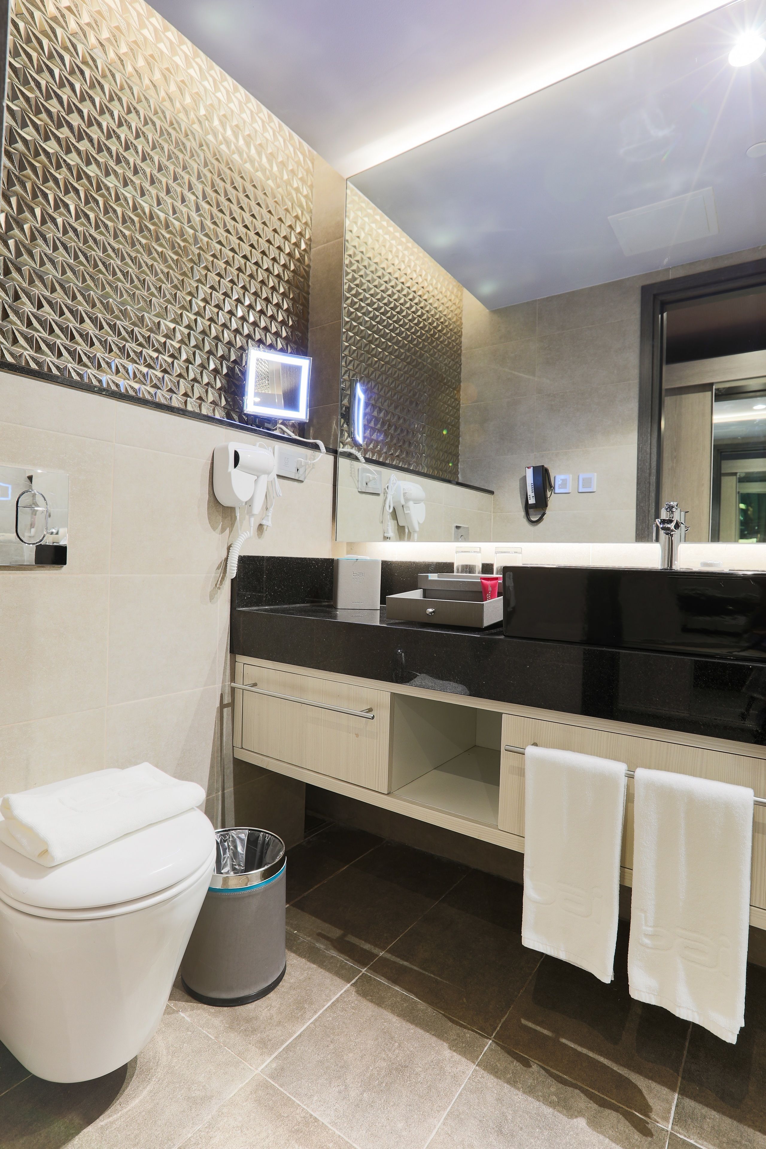 Un baño moderno con encimera negra, lavabo blanco y espejo sobre él. Hay un inodoro, un cubo de basura y dos toallas colgadas en la pared.
