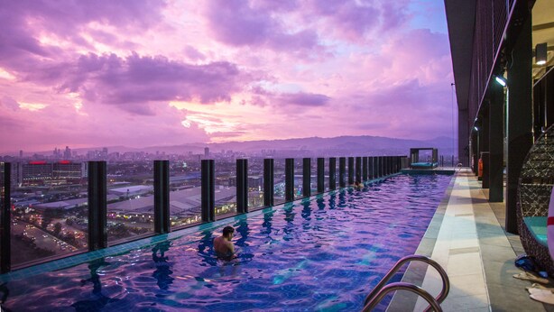 An infinity pool with a city view, a glass barrier, and a poolside area with a chair. - bai Hotel Cebu (Mandaue)