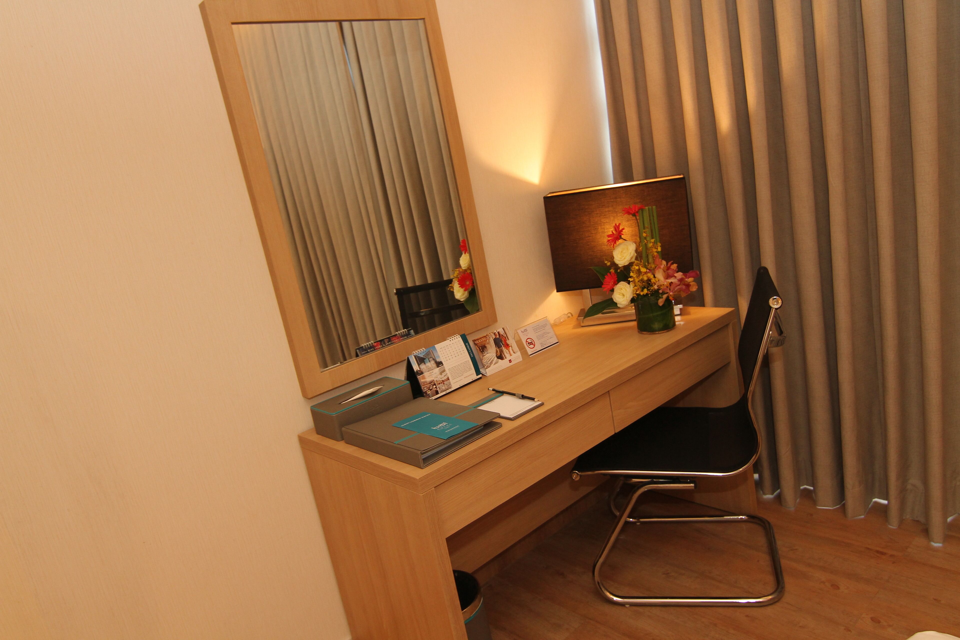 Habitación de hotel con escritorio de madera, una silla, un espejo, una lámpara y una ventana con cortinas.