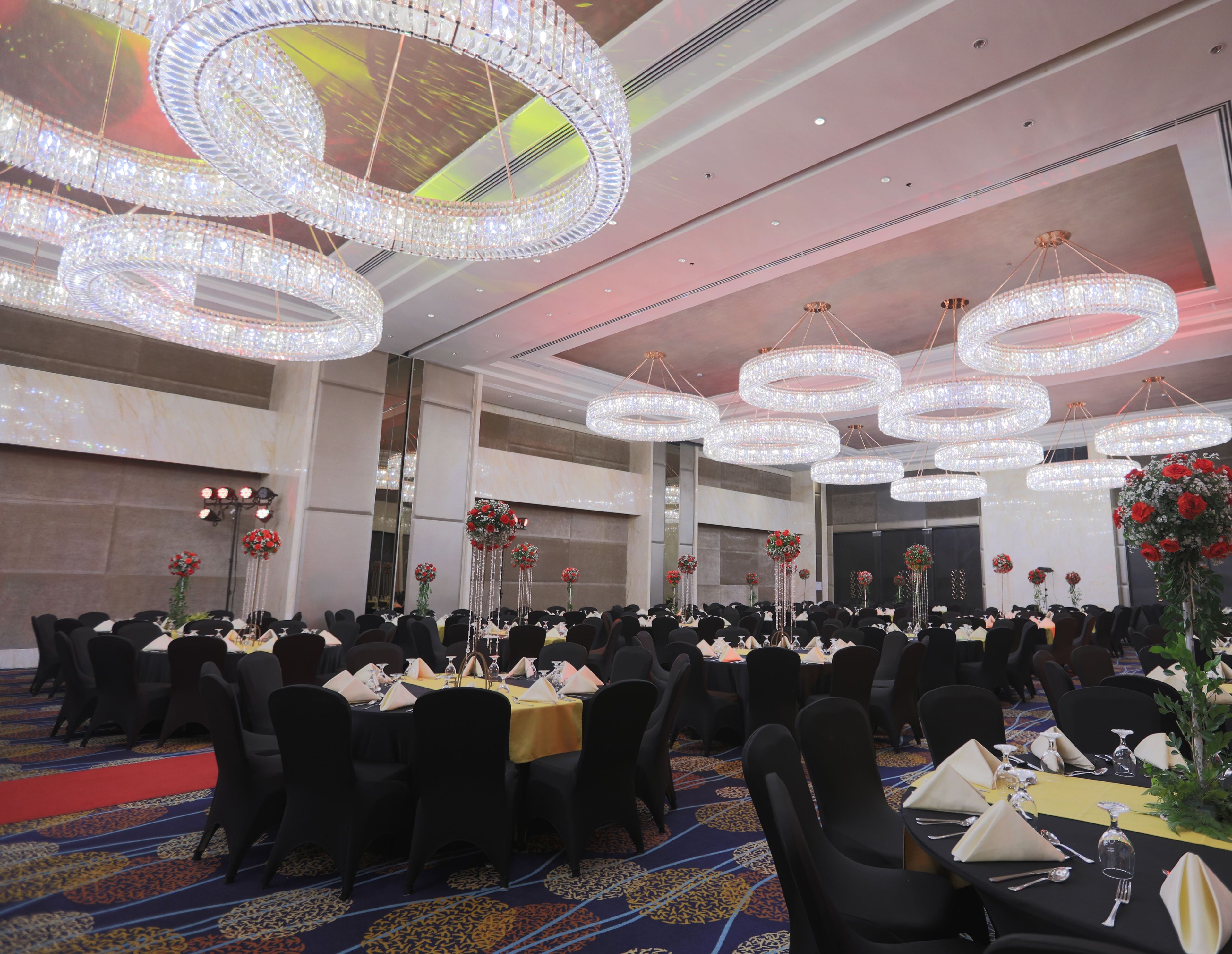 A banquet hall with multiple round chandeliers, tables set with white tablecloths and black chairs, and floral centerpieces.
