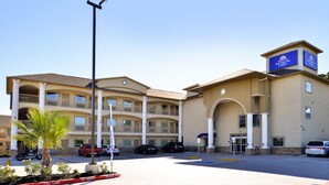 A multi-story hotel with a central entrance, parking area, and a palm tree in the foreground. - Americas Best Value Inn & Suites Spring Houston N (Spring)