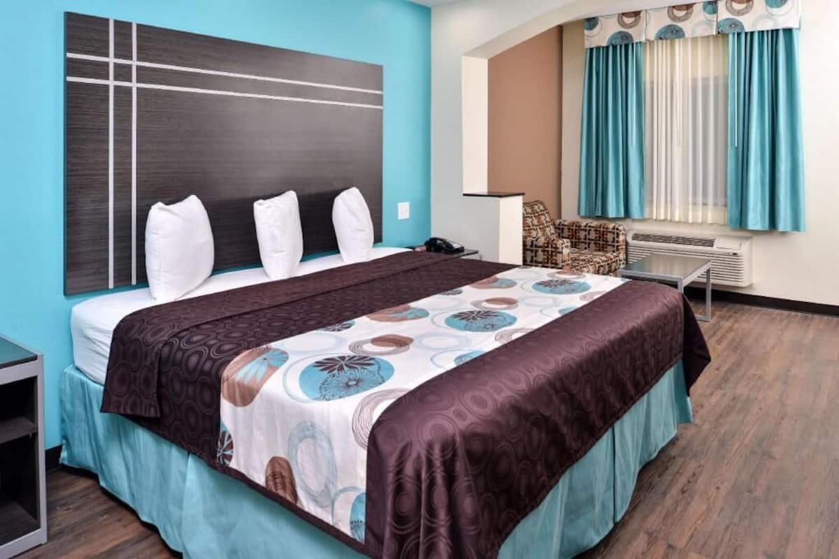 A hotel room with a bed, a bedside table, a chair, a window with curtains, and an air conditioning unit.