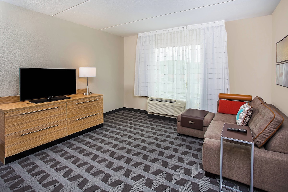 A hotel room with a flat-screen TV, a lamp, a sofa, a small table, and a window with curtains.