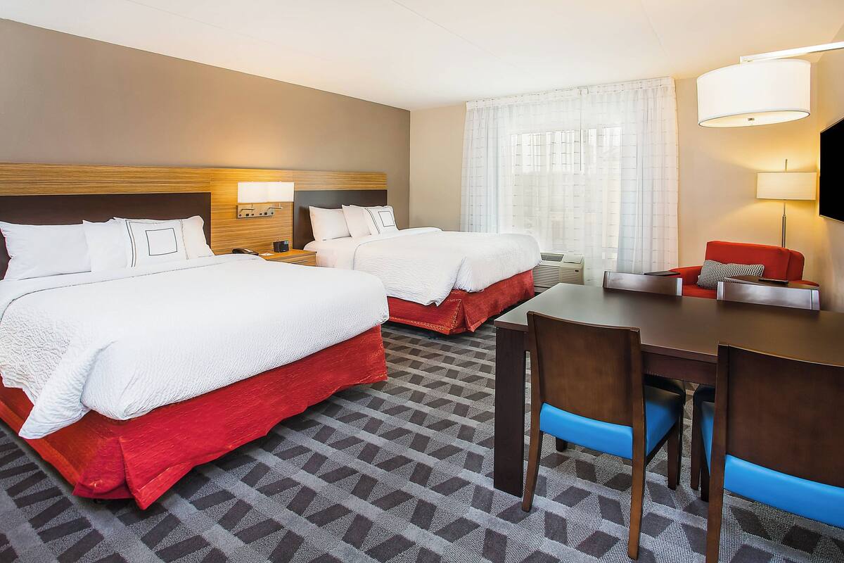 A hotel room with two beds, a desk, chairs, a TV, and a window with curtains.