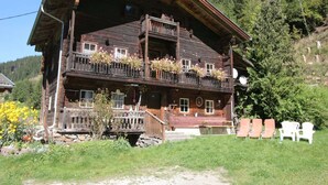 A traditional wooden chalet with a balcony, surrounded by greenery and a mountainous backdrop.