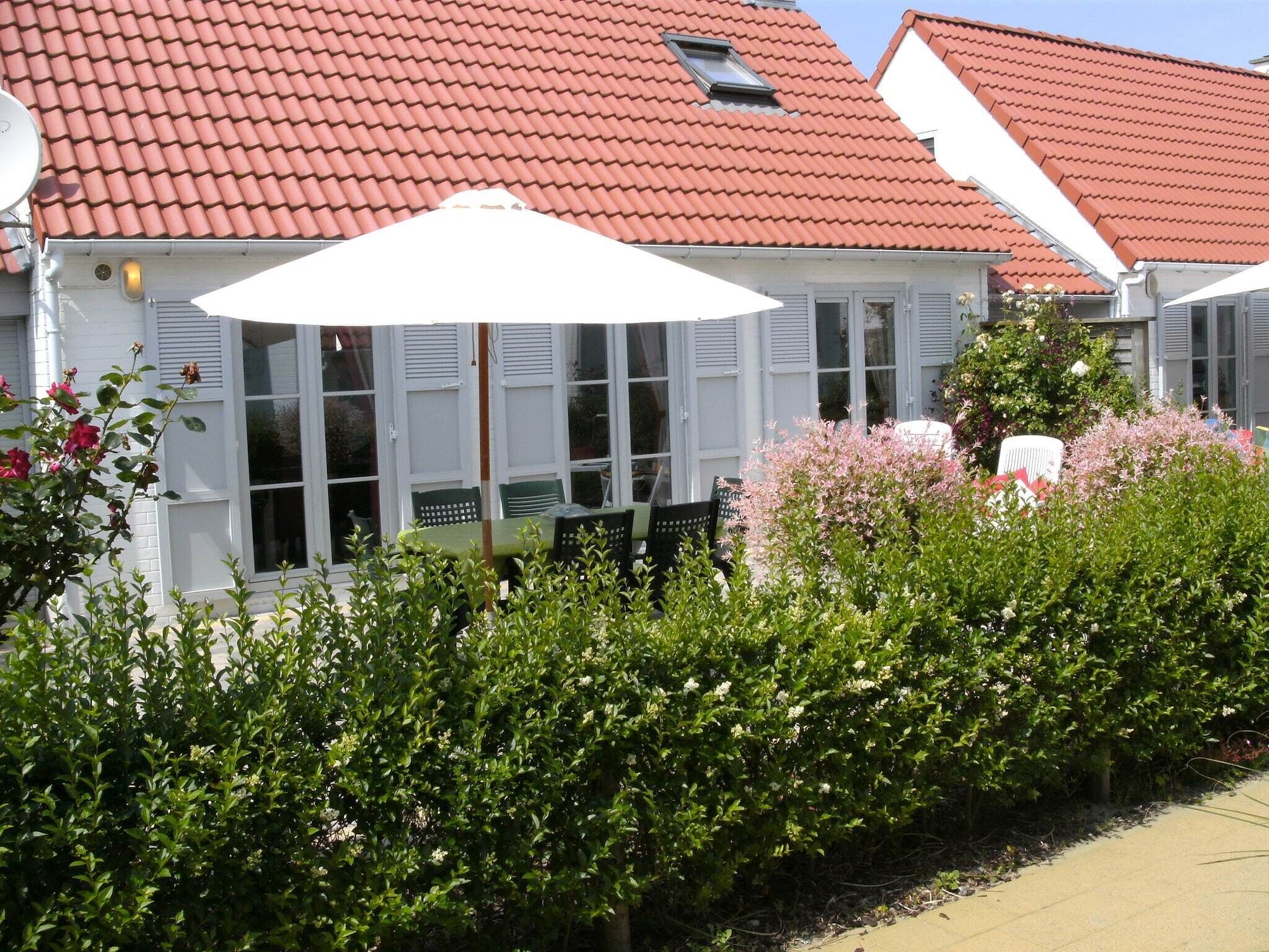 Ein weißes Gebäude mit rotem Ziegeldach, einer überdachten Außenbereich-Sitzgelegenheit mit Tischen und Stühlen sowie einem gepflegten Garten mit Sträuchern und Blumen.