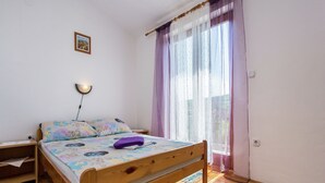Une chambre à coucher avec un lit en bois, une table de chevet, une lampe, une fenêtre avec des rideaux légers et une vue sur l’extérieur.