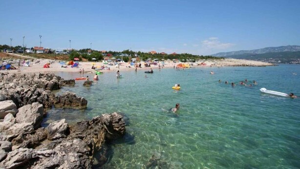 Une plage à l’eau claire, avec un rivage rocheux, où l’on peut voir des gens nager et prendre des bains de soleil.