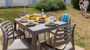 Mesa de refeições ao ar livre com cadeiras, guarda-sol e comida, incluindo pão, fruta e bebidas.