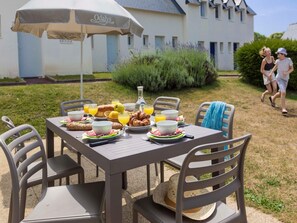 Ein Essplatz im Freien mit Tisch, Stühlen und Sonnenschirm, gedeckt mit Brot, Obst und Getränken.
