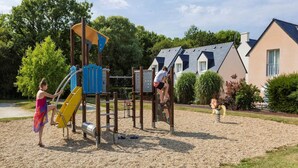 Um parquinho infantil com estruturas de escalada e um escorrega, cercado por uma área de brita e edifícios residenciais.