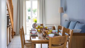 Uma sala de jantar com uma mesa de madeira posta para o café da manhã, cadeiras, uma luminária pendente e vista para o exterior através de uma porta de vidro.