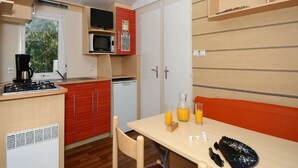 A compact kitchen with a gas stove, microwave, and a small refrigerator. There is a dining table with a glass of orange juice and a bottle. - Cap d’Agde Bungalow Retreat (Cap D'agde)