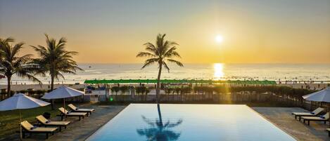 Ein Pool mit Liegestühlen und Sonnenschirmen, ein Strand mit Menschen und ein Sonnenuntergang über dem Meer.