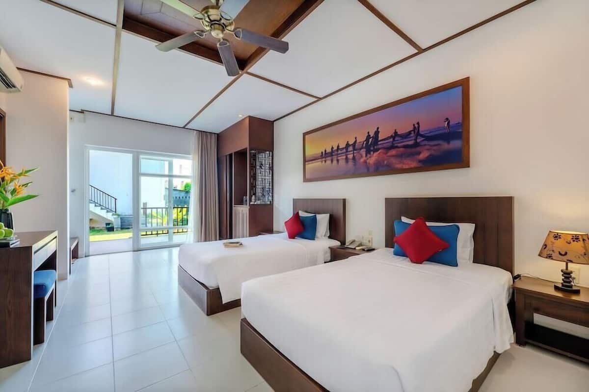 A hotel room with two beds, a ceiling fan, a painting, a desk, and a view of the outdoors.