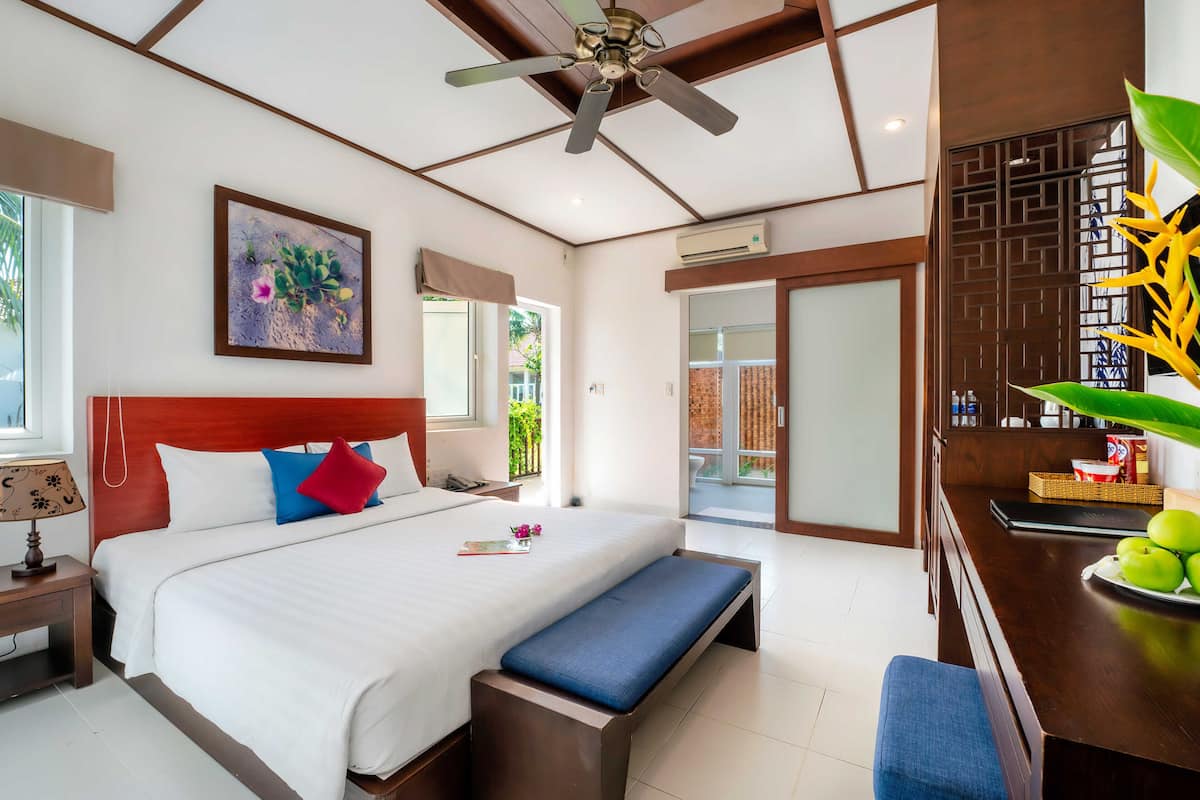 A hotel room with a bed, a desk with a laptop, a ceiling fan, and a view of the outside.