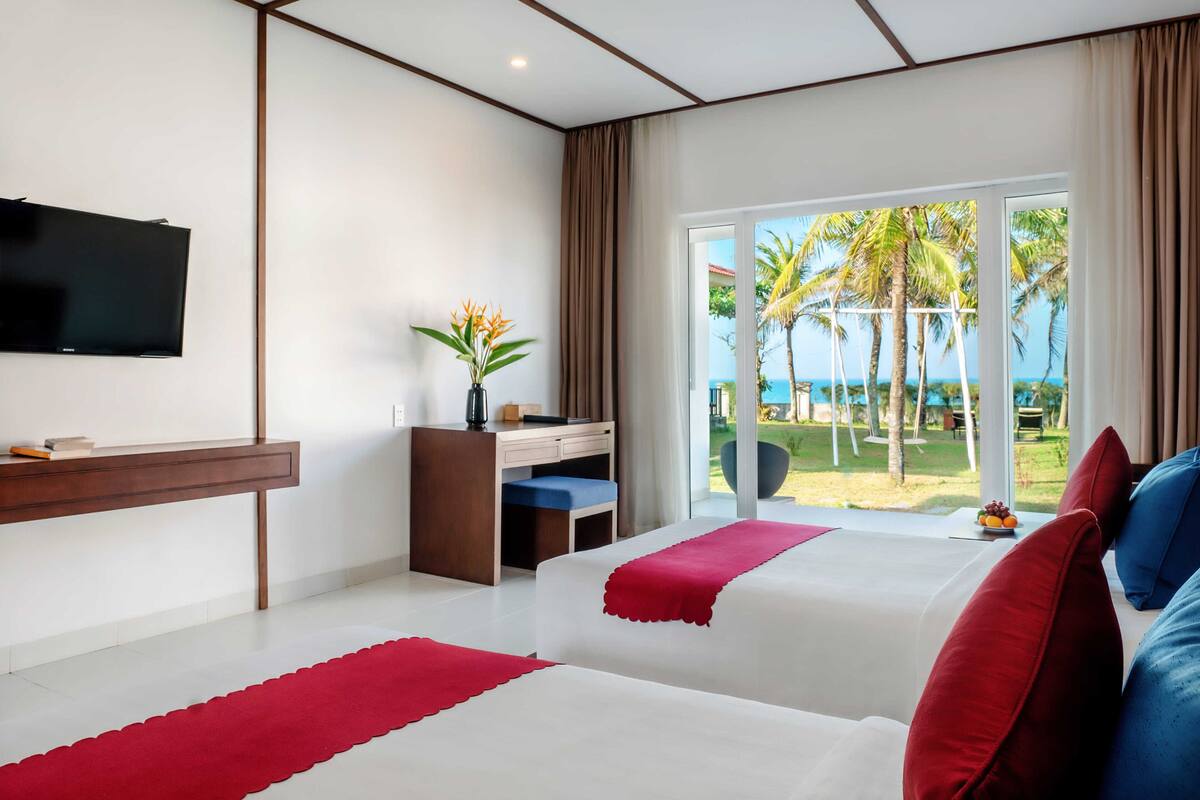 A hotel room with two beds, a desk, a TV, and a view of the beach.