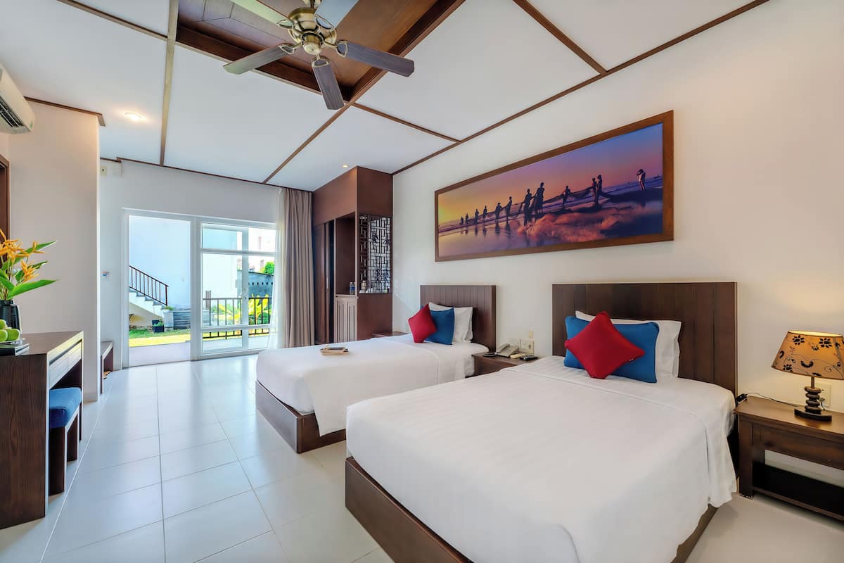 A hotel room with two beds, a desk, a chair, a ceiling fan, a painting, and a view of the outdoors.