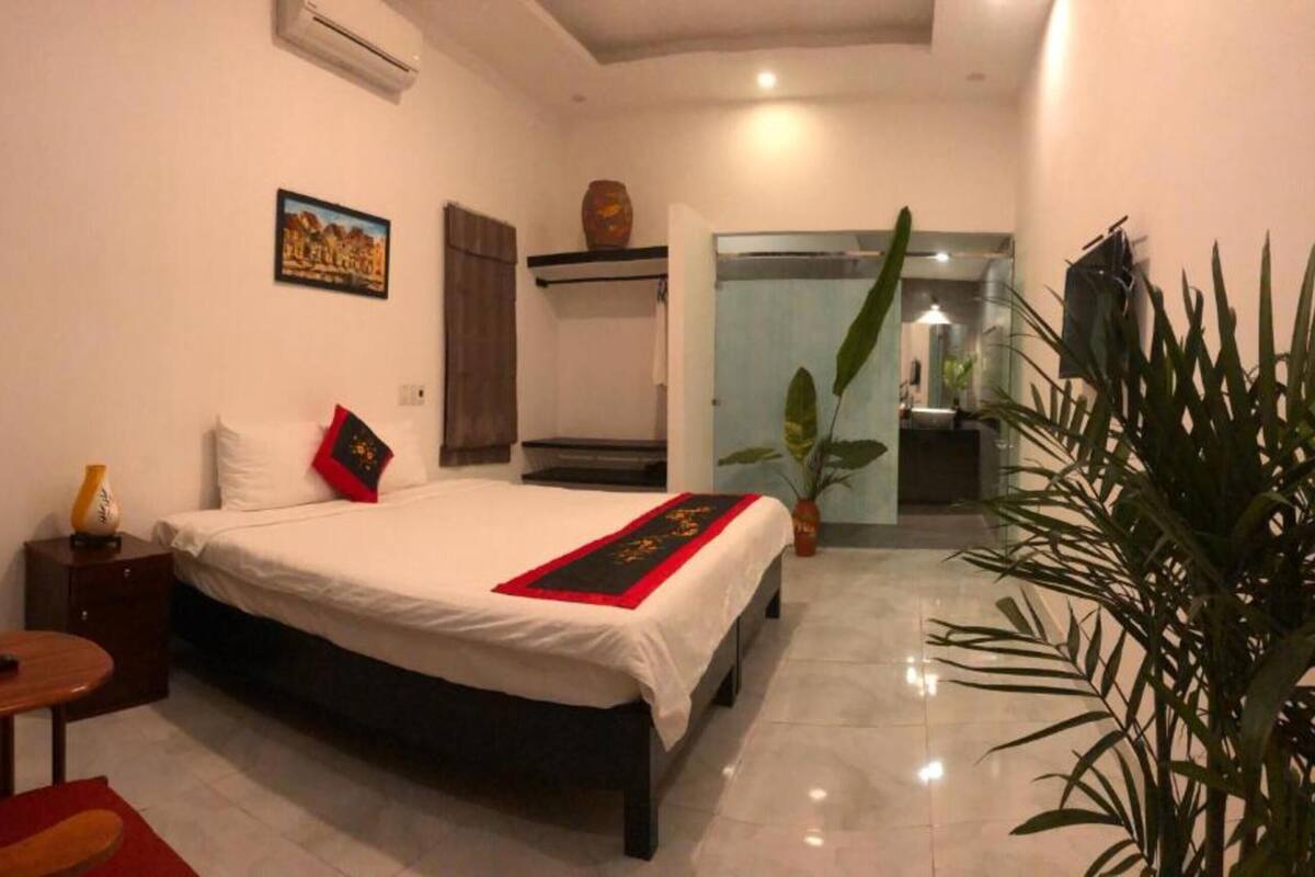 Un dormitorio con cama, mesita de noche, una silla, una pequeña mesa, una planta y un baño visible a través de una puerta abierta.