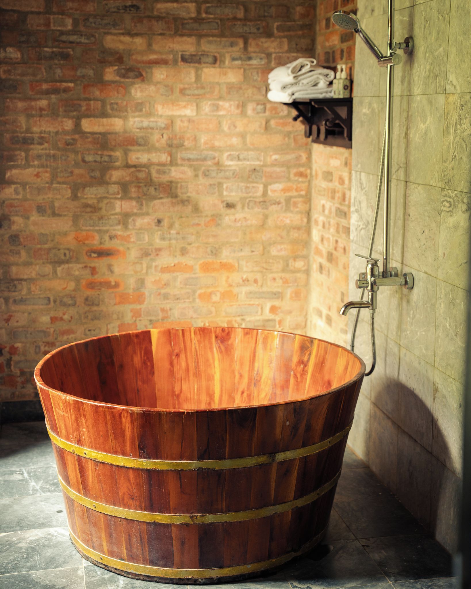 A wooden barrel bathtub in a bathroom with a brick wall and a shower.