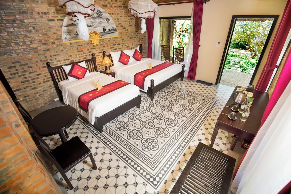 A room with two beds, a small table, a chair, and a view of greenery outside.