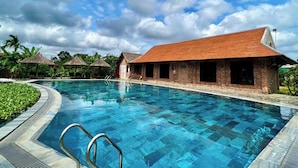 A swimming pool with a clear blue water surface, surrounded by a brick building with a red roof, and a tropical setting with palm trees. - Hue Ecolodge (Hue)