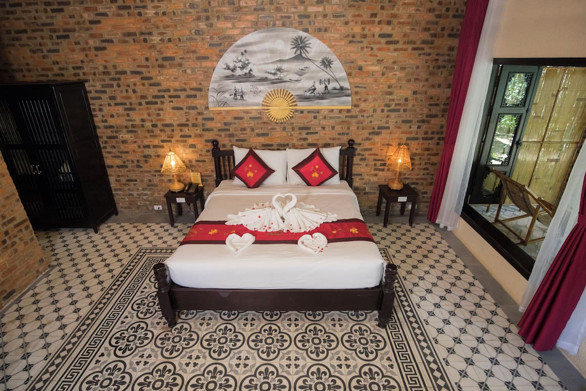 A neatly made bed with decorative swans and red pillows, a brick wall with a mural, and a patterned floor.