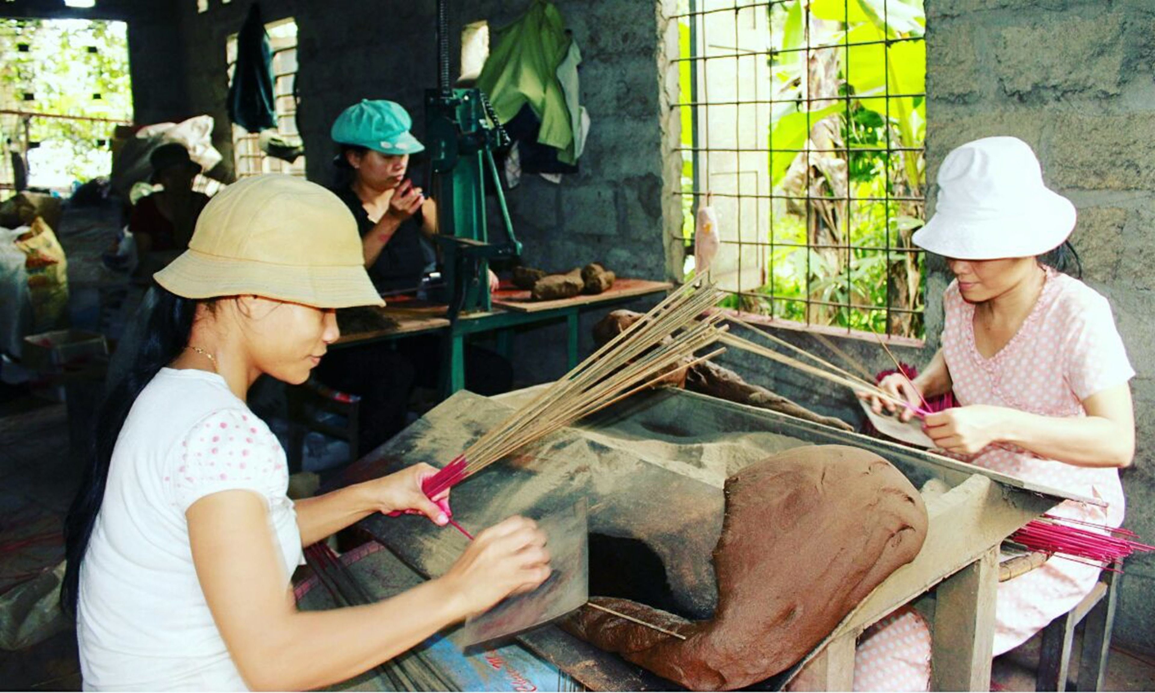 A workshop with individuals engaged in crafting activities, featuring a workbench, tools, and a window with greenery outside.
