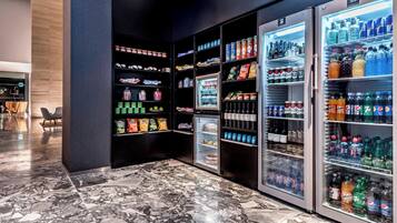 A modern hotel lobby with marble flooring, a seating area with chairs, and a row of beverage vending machines stocked with various drinks and snacks.